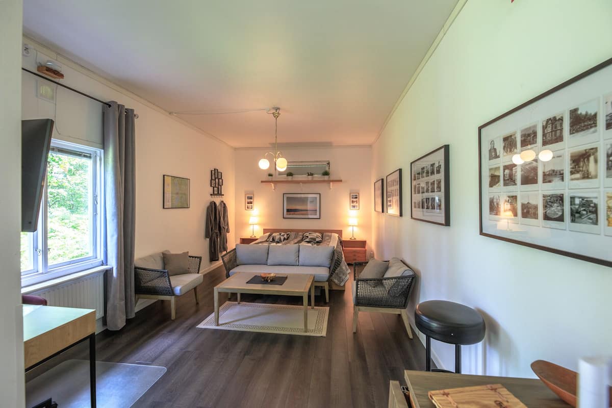 Ein kompakter Wohnbereich mit Bett, Sofa und Essbereich. An der Wand hängen gerahmte Bilder, ein Fenster mit Vorhängen und ein Deckenventilator.
