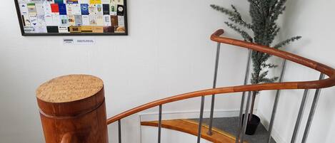 Una escalera de madera con barandilla de corcho, un collage enmarcado de tarjetas de presentación en la pared, y un pequeño árbol artificial a su lado.