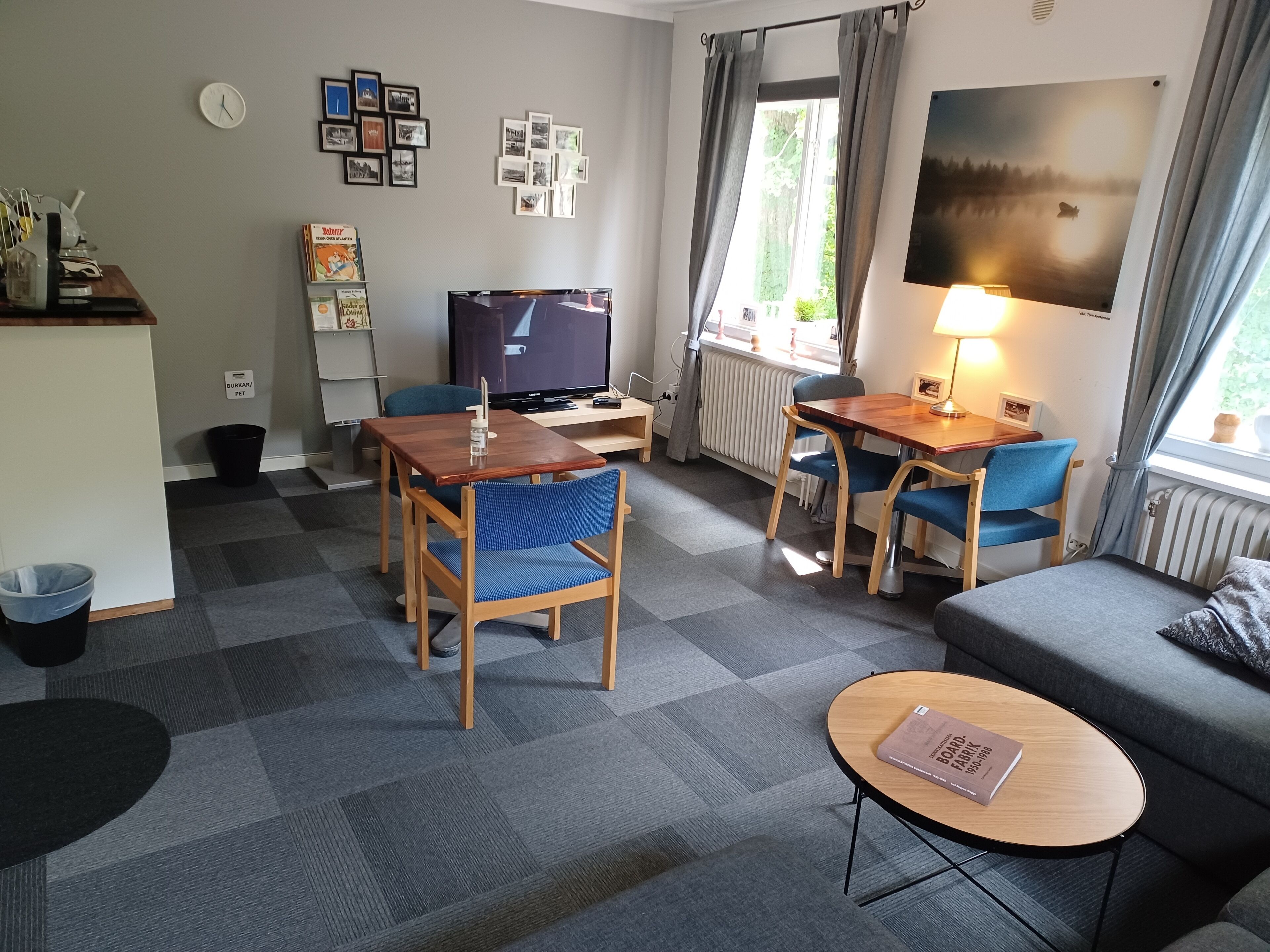 Une chambre d’hôtel moderne avec un canapé, une table à manger avec des chaises, une télévision, une petite table avec un livre et une fenêtre avec des rideaux.