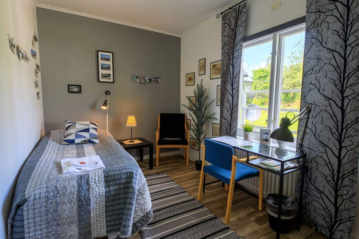 Ein Zimmer mit Bett, Schreibtisch, Stuhl, Lampe, einem Fenster mit Vorhängen und einer kleinen Pflanze.