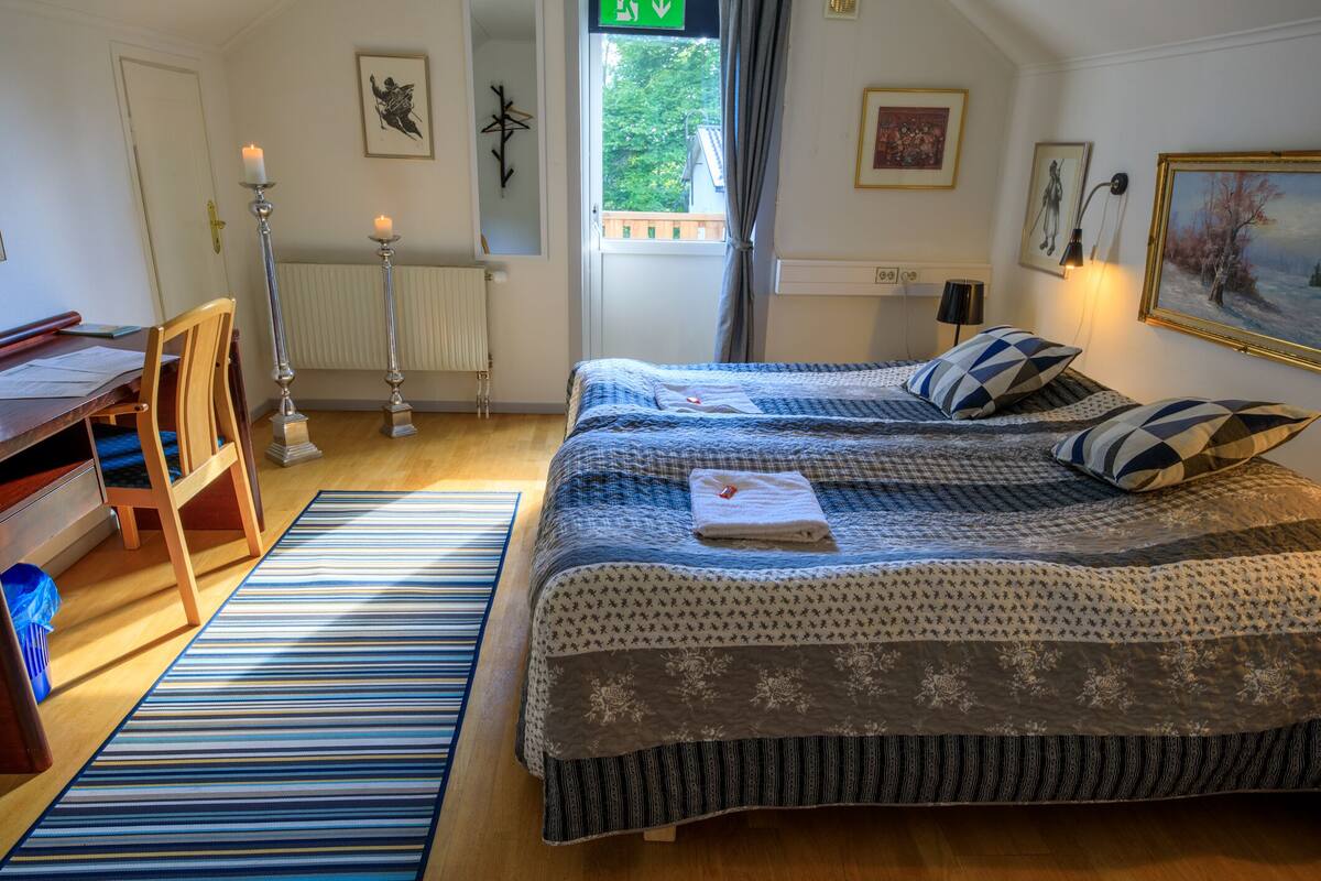 Ein Schlafzimmer mit zwei Betten, einem Holztisch, einem Heizkörper und einem Fenster mit Blick auf Grünflächen.