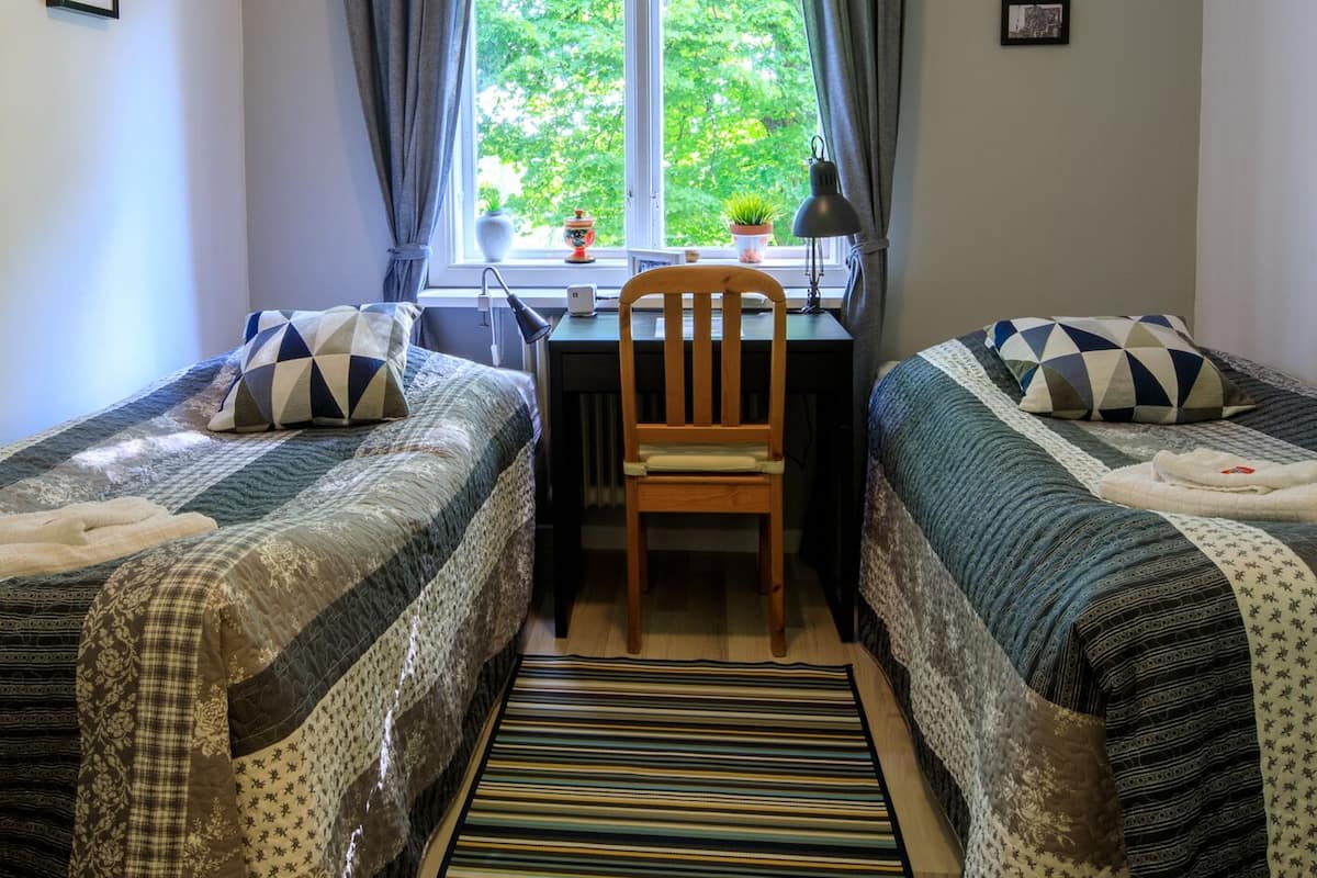 Ein Zimmer mit zwei Einzelbetten, einem Schreibtisch, einem Stuhl, einem Fenster mit Vorhängen und Blick ins Grüne.