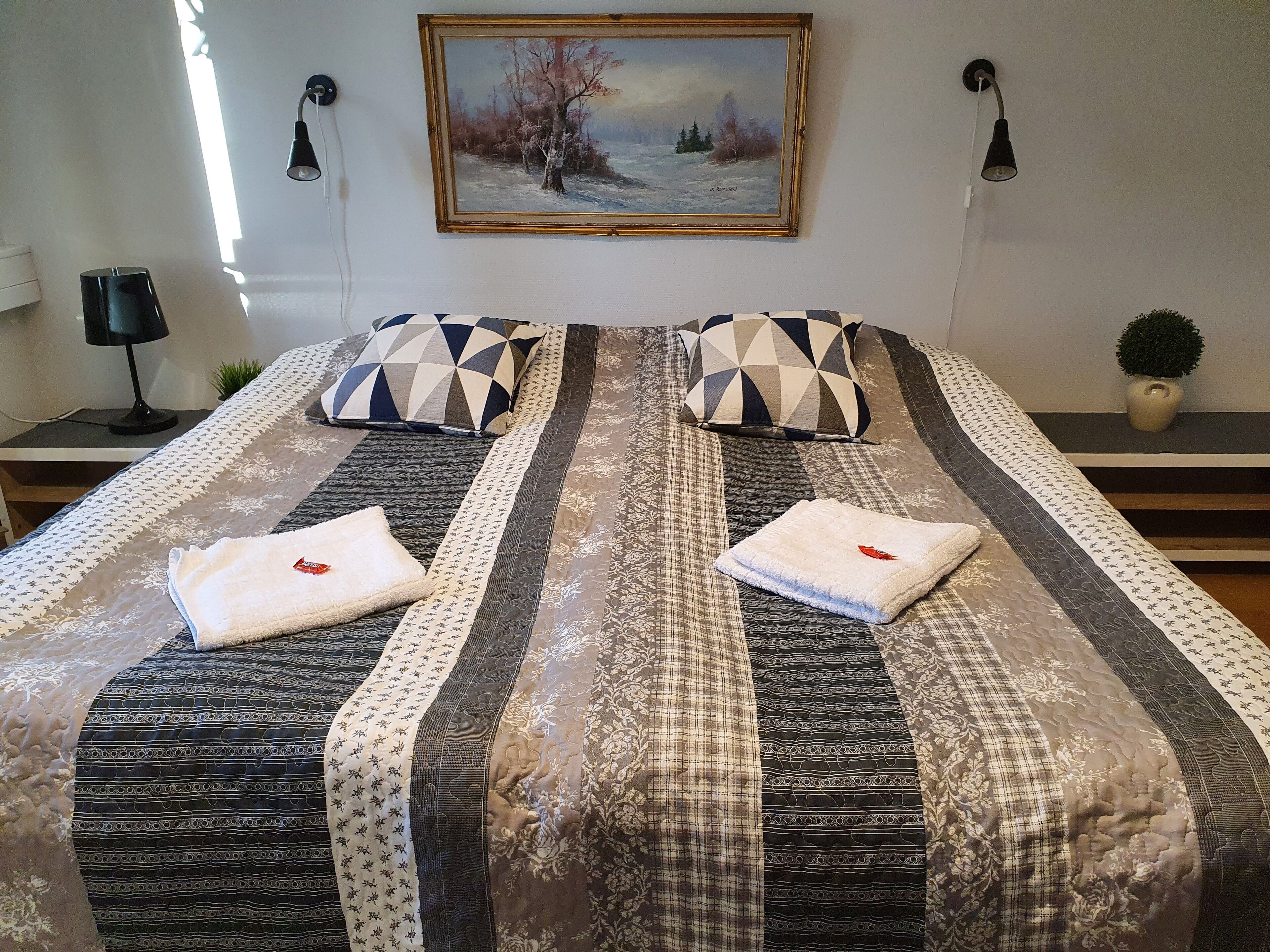 Una cama bien tendida con ropa de cama estampada y dos almohadas, un cuadro en la pared y una planta pequeña en la mesita de noche.