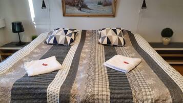Una cama bien tendida con ropa de cama estampada y dos almohadas, un cuadro en la pared y una planta pequeña en la mesita de noche.