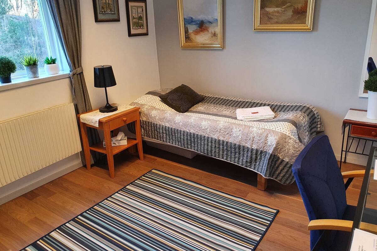 Ein ordentlich eingerichtetes Schlafzimmer mit Bett, Nachttisch, Lampe und Blick ins Freie durch ein Fenster.