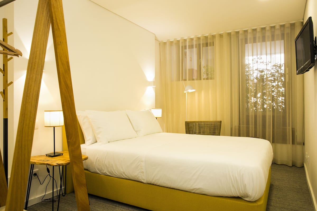 Hotelkamer met een bed, nachtkastje met lamp, stoel, televisie aan de muur en dunne gordijnen.