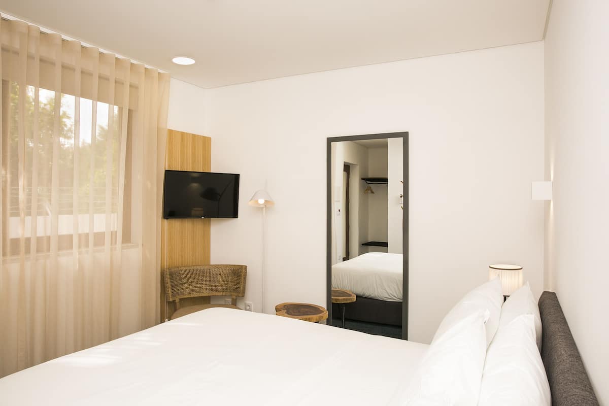 Een hotelkamer met een bed, een aan de muur bevestigde televisie, een spiegel en een raam met dunne gordijnen.