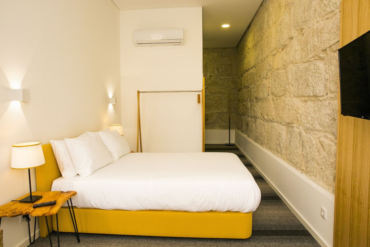 Een moderne hotelkamer met een geel bed, witte lakens, een houten nachtkastje met een lamp, en een stenen muur.