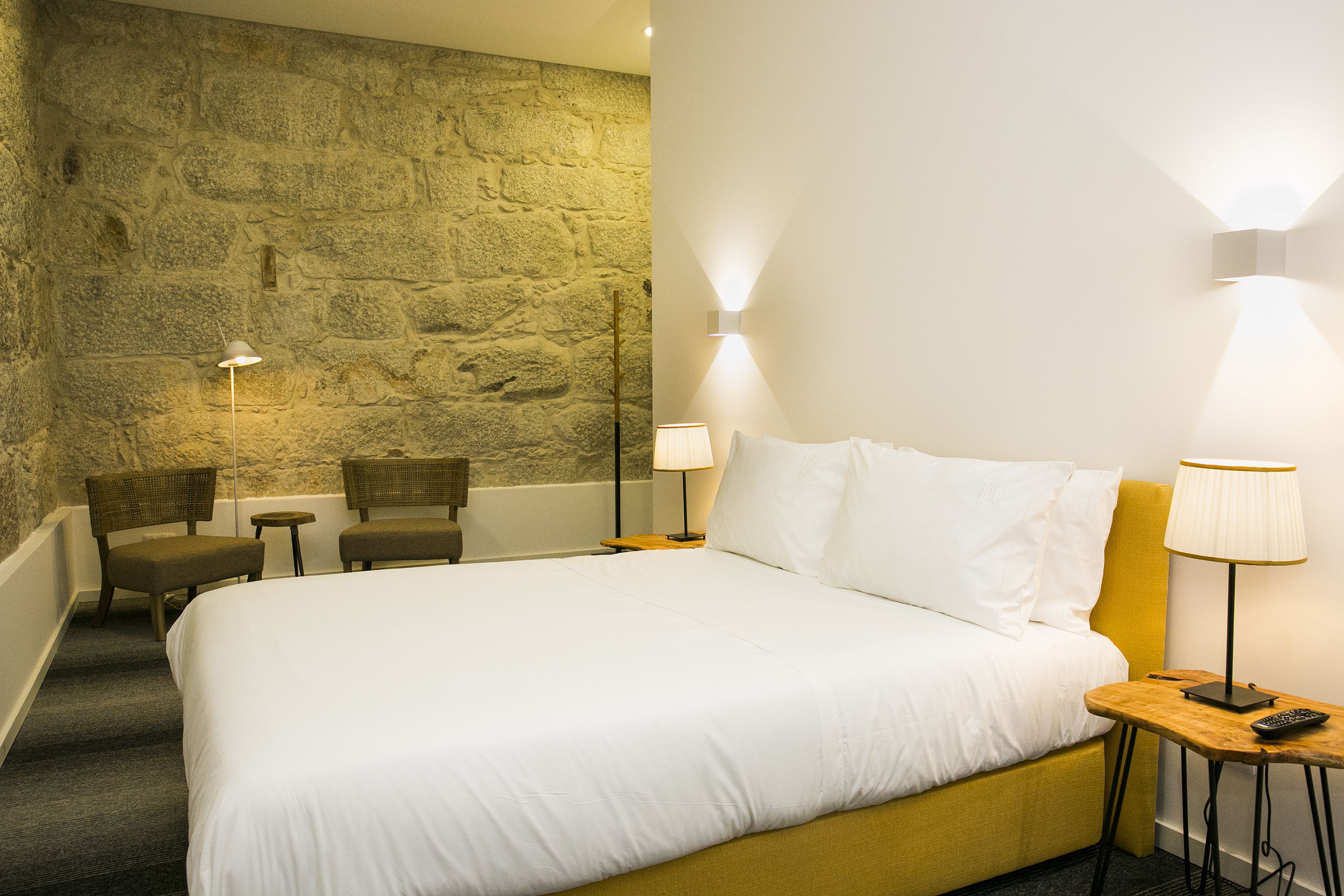 Une chambre d’hôtel avec un grand lit, deux chaises, une petite table et un mur en pierre.
