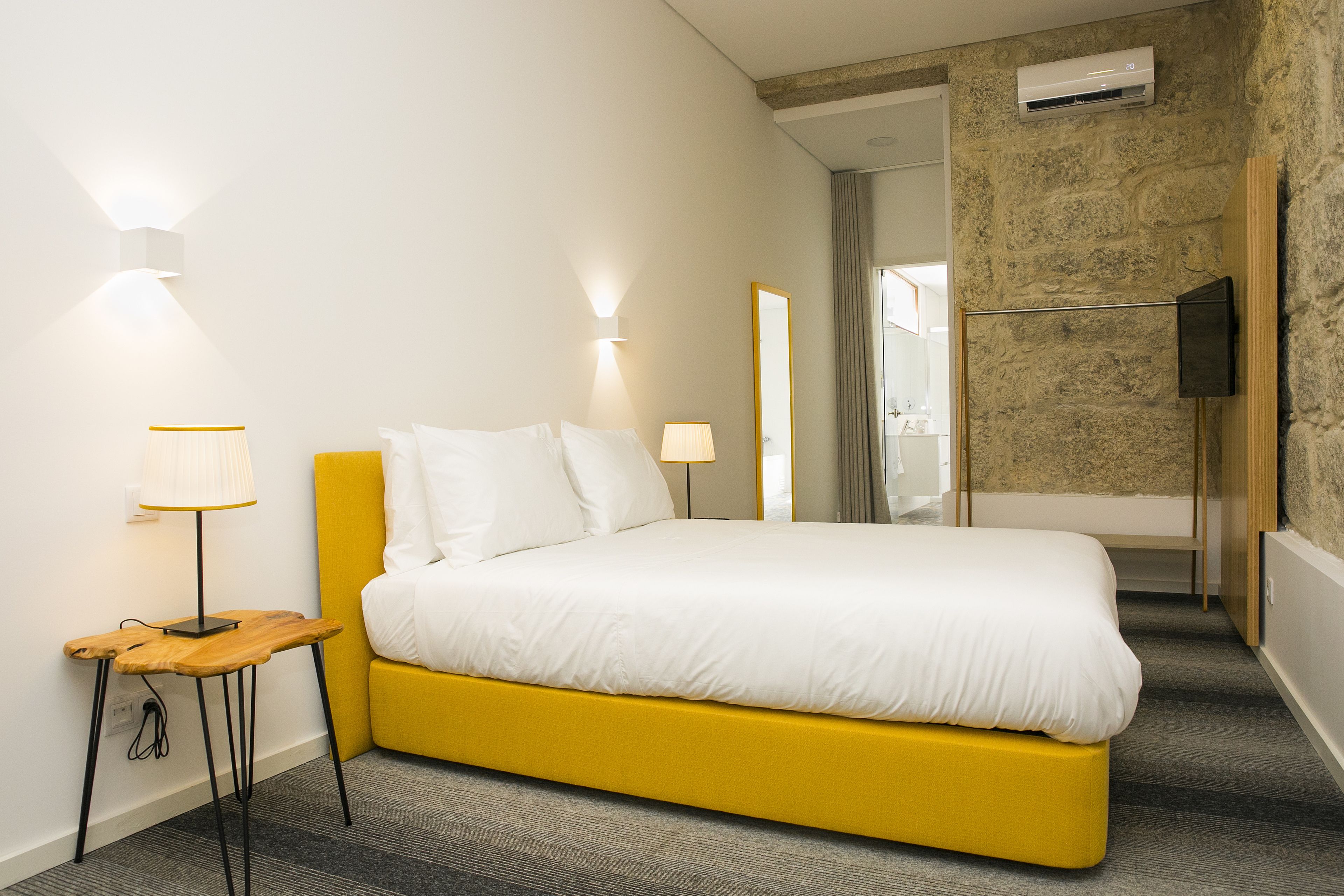 Habitación de hotel moderna con una cama amarilla, una mesita de noche pequeña con una lámpara, un televisor montado en la pared y un equipo de aire acondicionado.