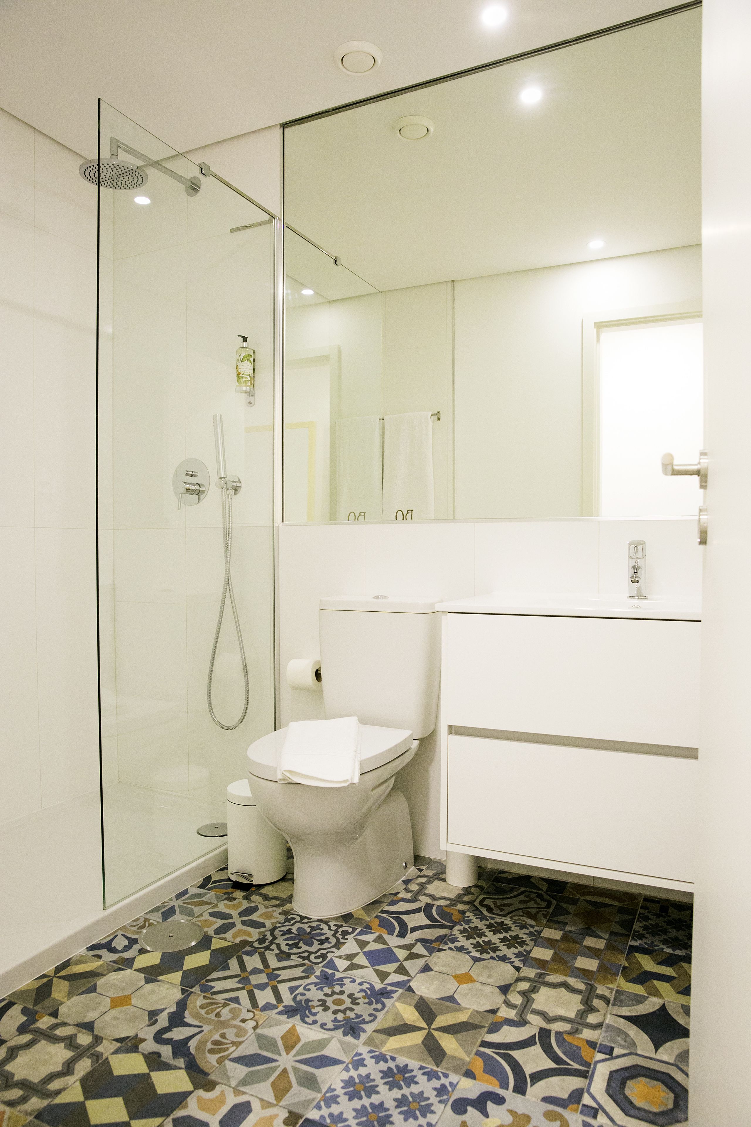 Een moderne badkamer met een douchecabine van glas, een toilet en een wastafel met een spiegel erboven. De vloer heeft een betegeld patroon.