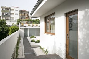 En modern bostadsbyggnad med en smal gång som leder till en dörr, omgiven av grönska och en vit vägg.