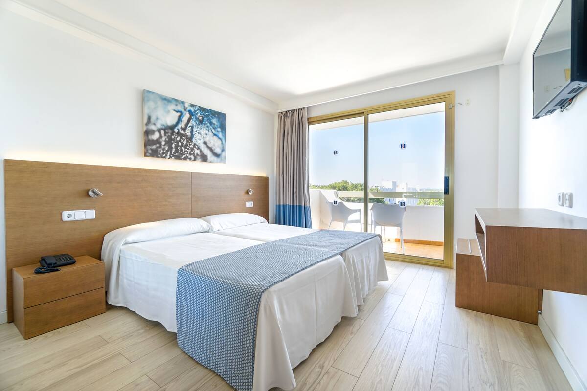A hotel room with a bed, bedside table, and a desk. There is a large window with a view of the city.
