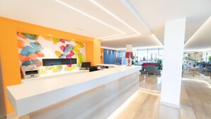 Un espace d’accueil moderne, doté d’un comptoir de réception blanc, d’œuvres d’art murales abstraites et colorées, et d’un aménagement spacieux avec une grande fenêtre laissant entrer la lumière naturelle.
