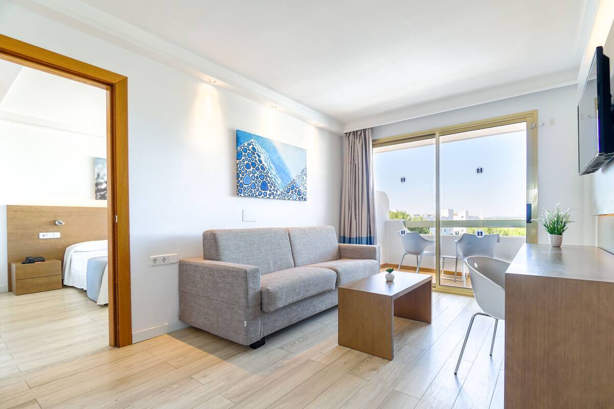 A modern hotel room with a sofa, a small table, a balcony with a view, and a TV mounted on the wall.