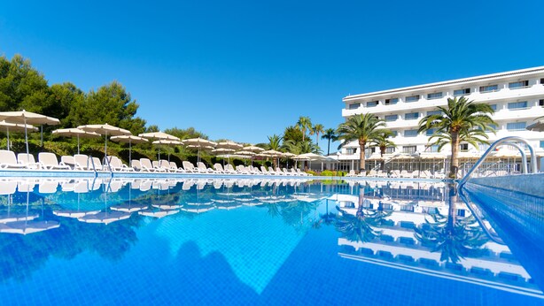 Une piscine entourée de palmiers et d’un bâtiment d’hôtel, avec des chaises longues et des parasols à proximité.