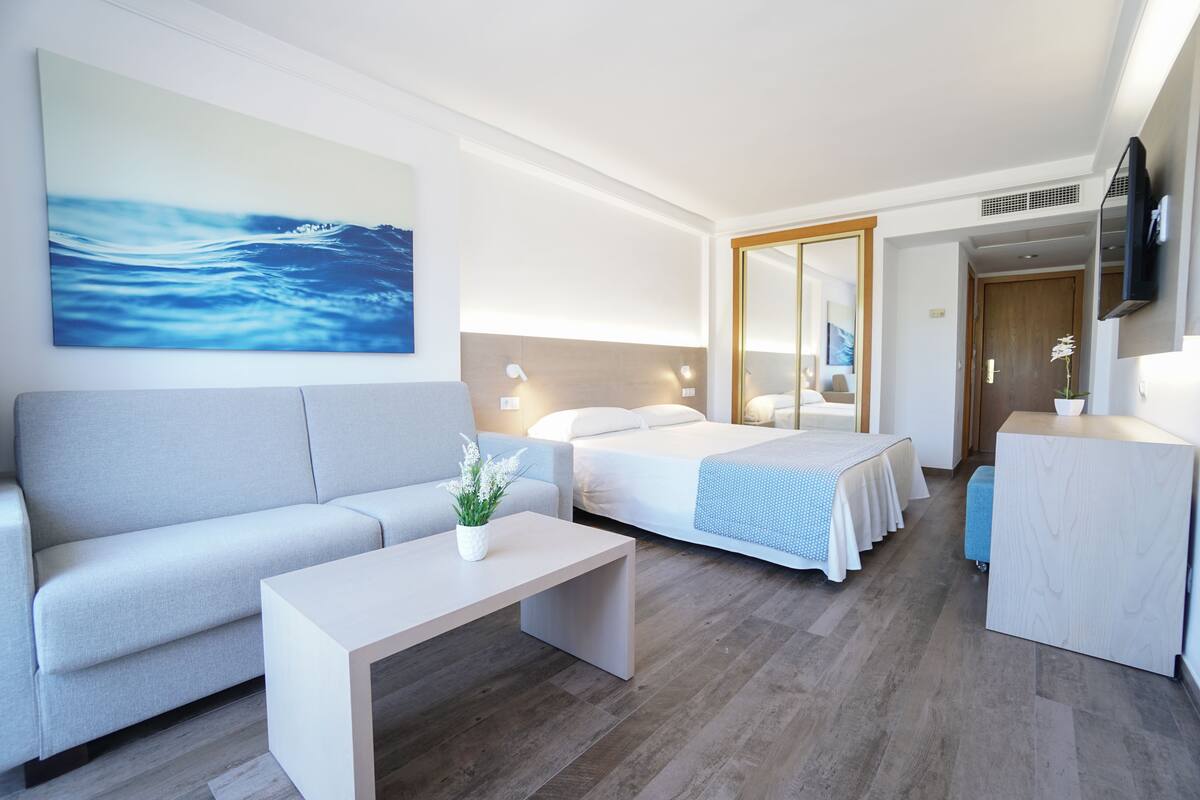 A modern hotel room with a sofa, a bed with a blue bedspread, a bedside table with a plant, and a large painting of a wave on the wall.