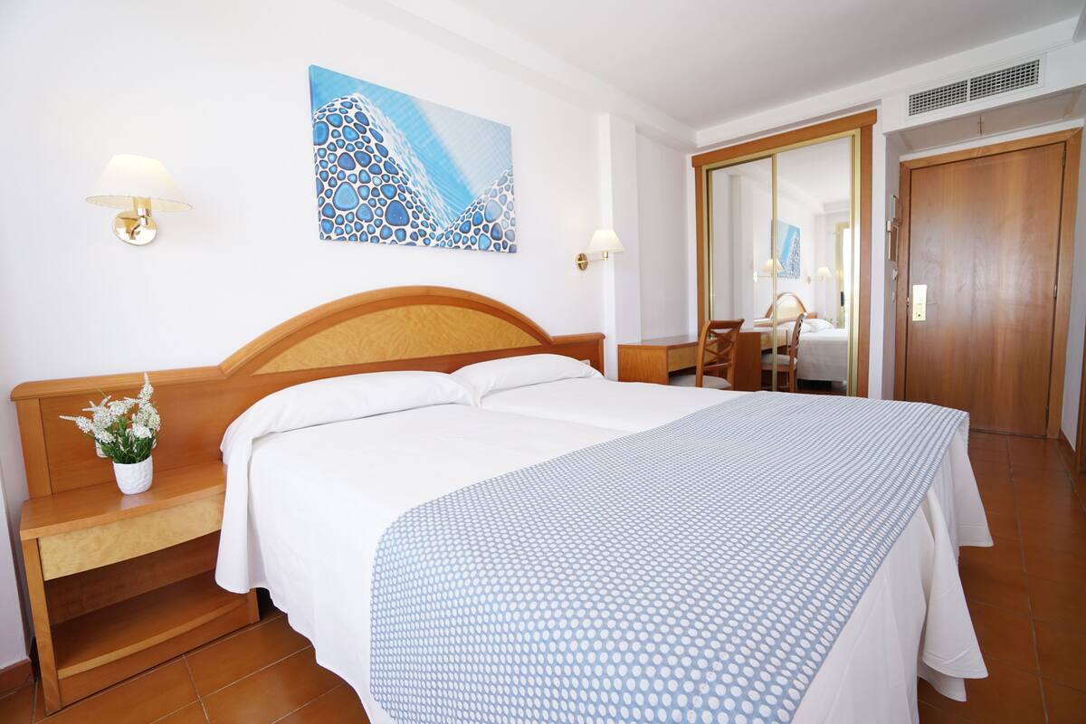 A hotel room with a wooden bed, a bedside table with a lamp, a mirror, and a painting on the wall.
