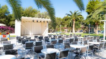 An outdoor seating area with multiple tables and chairs, surrounded by palm trees and greenery.