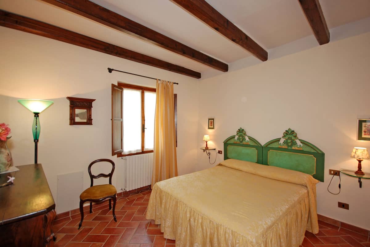 A bedroom with a bed, a chair, a table, a lamp, a window with curtains, and wooden beams on the ceiling.