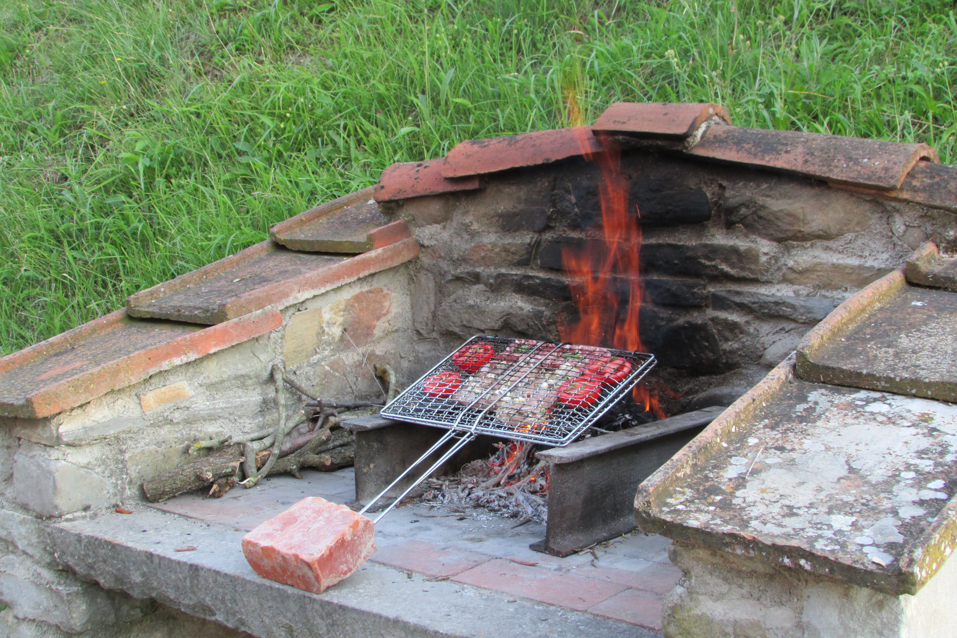 Un barbecue traditionnel en pierre, installé à l’extérieur, avec une grille en métal, entouré d’escaliers en pierre et d’une zone herbeuse.