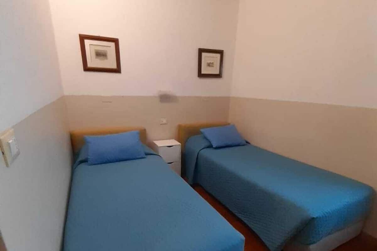 Une petite chambre propre, avec deux lits simples, une table de chevet et deux tableaux encadrés au mur.