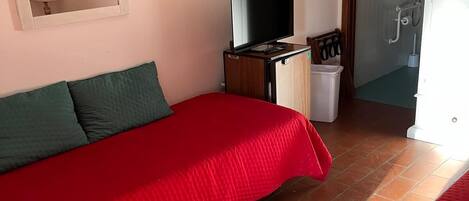 A bedroom with a red bed, a television, a small cabinet, and a bathroom visible through an open door.