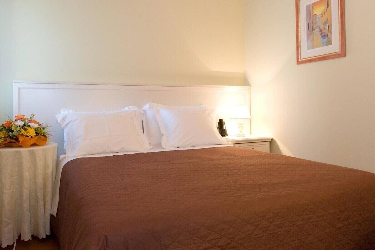 Un lit bien fait, avec une literie blanche et marron, une petite table sur laquelle est posé un vase de fleurs et un tableau encadré accroché au mur.