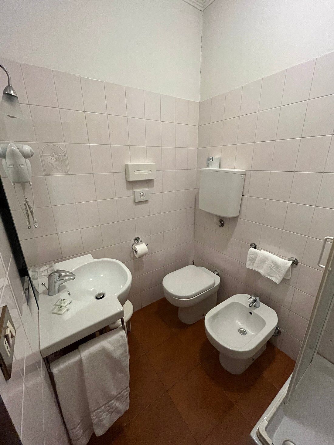 A small, modern bathroom with white tiles, a sink, a toilet, a bidet, and a shower.