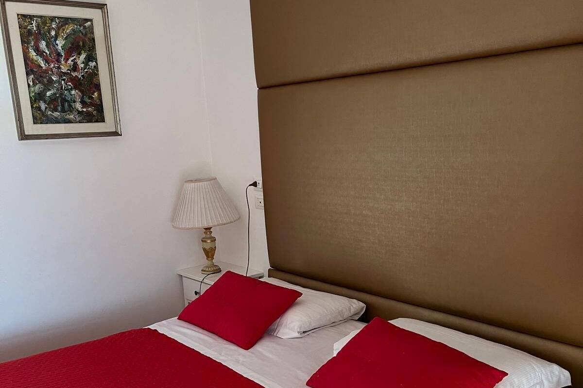 Un lit bien fait, avec une literie rouge et blanche, une tête de lit beige, une table de chevet blanche avec une lampe, et un tableau encadré au mur.