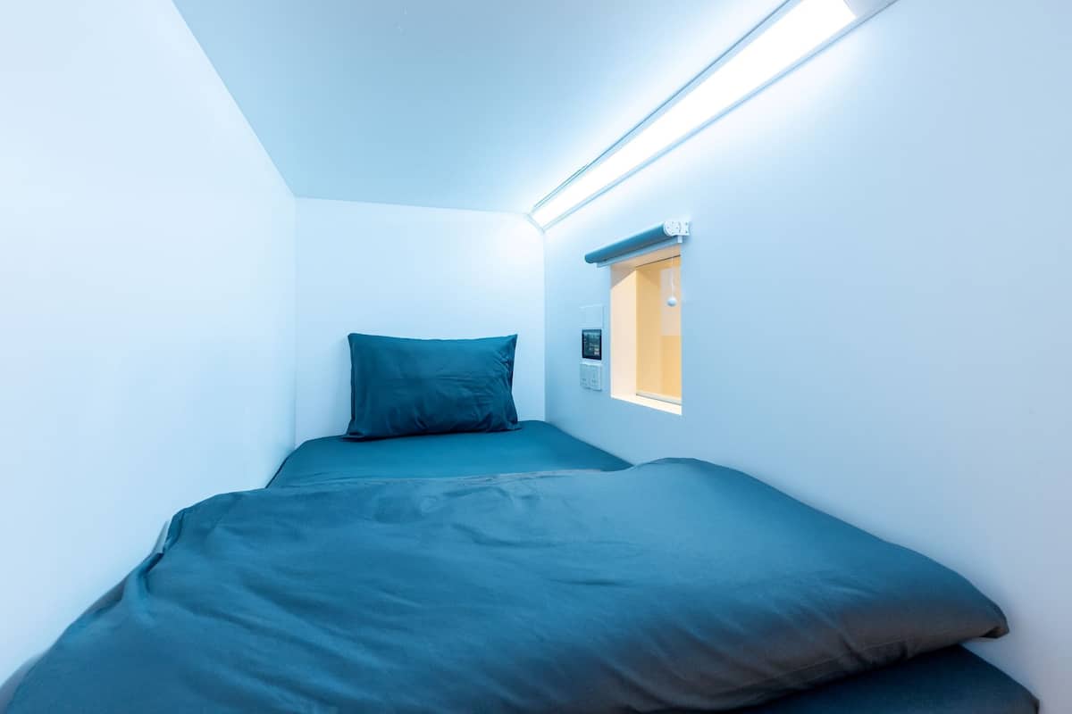 A small, narrow room with a bed, a window, and a ceiling light.