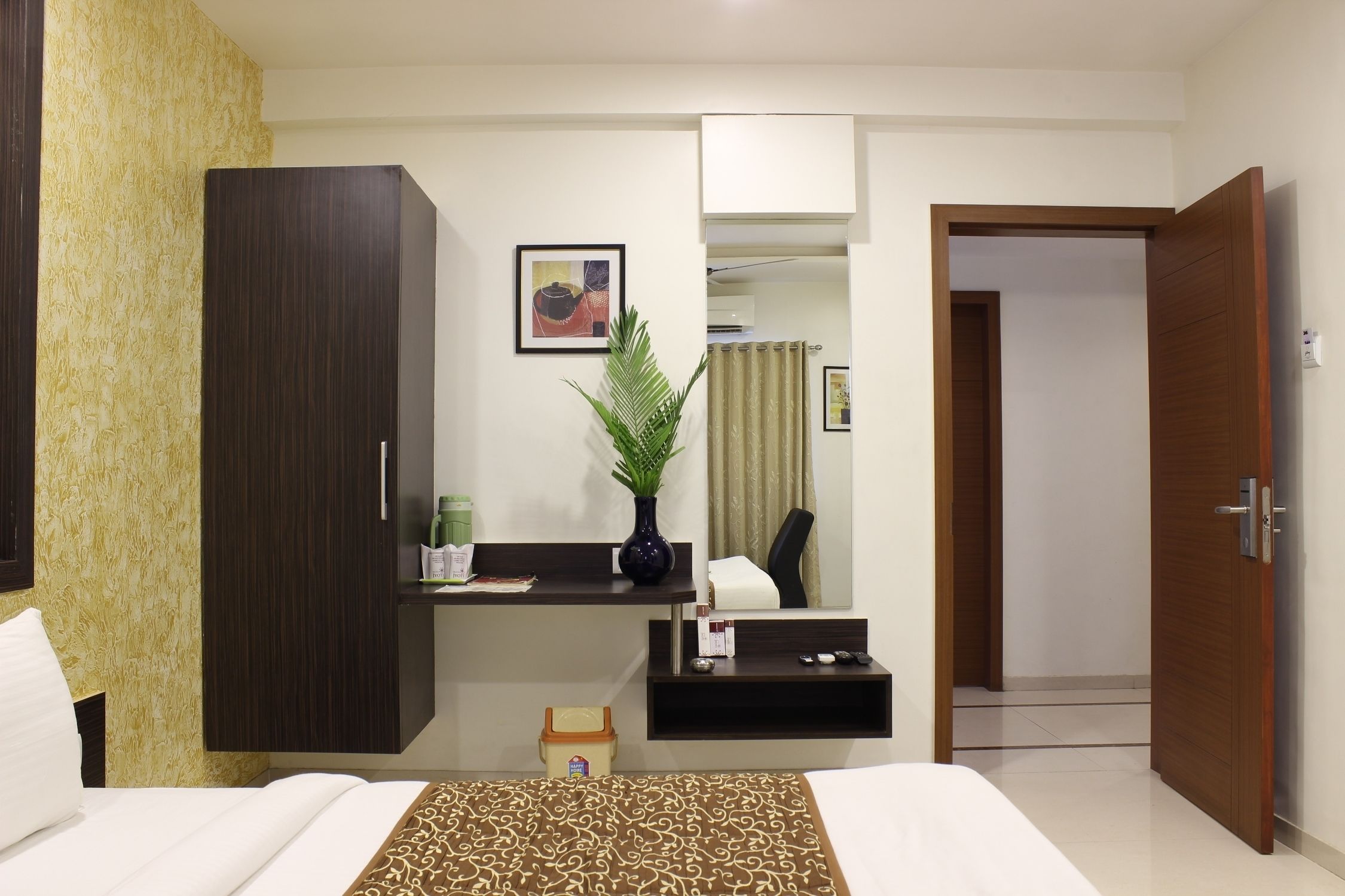 A hotel room with a bed, a desk, a wardrobe, a potted plant, and a bathroom door.