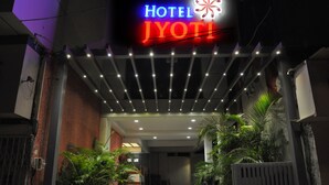 Entrada de un hotel con un letrero de neón que dice "Hotel Jyoti", rodeada de plantas en macetas y un corredor cubierto.