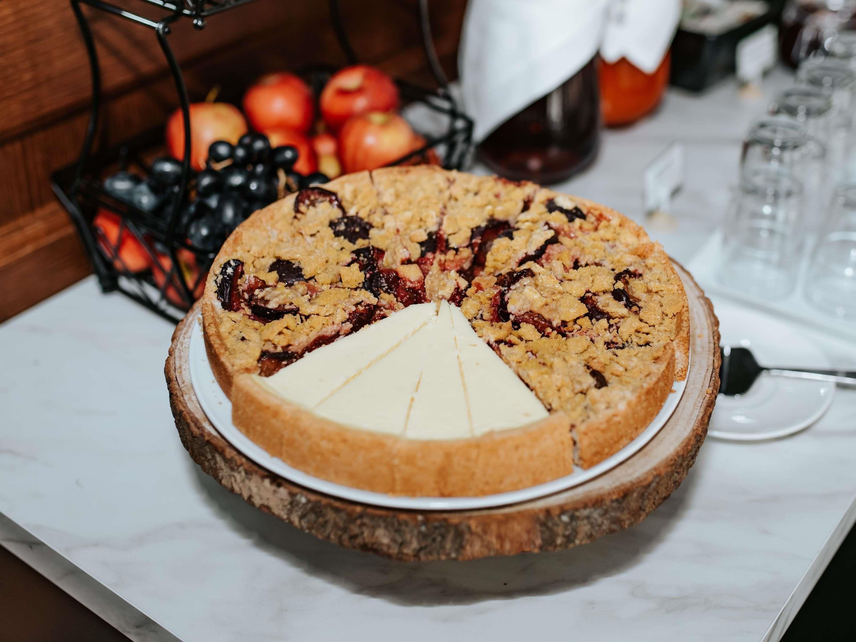A round pie with a crumbly topping on a wooden board, a slice cut out, and a fruit basket in the background.