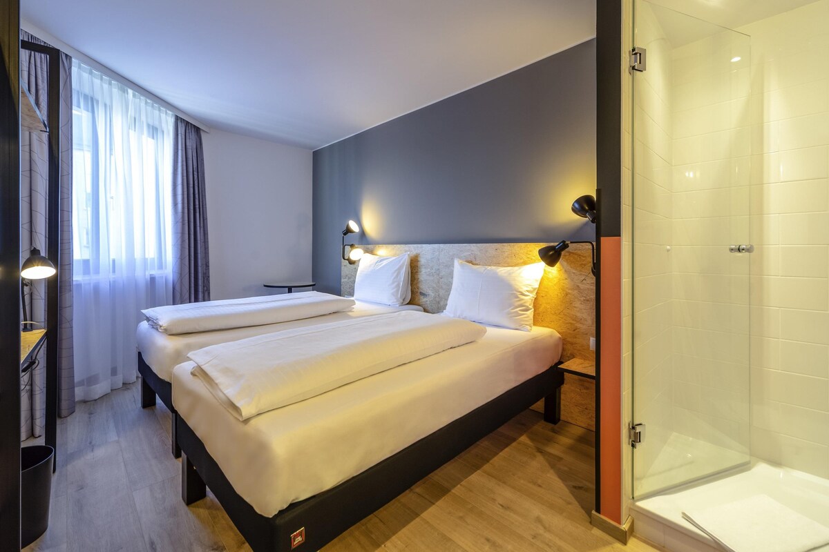 Una habitación de hotel moderna con dos camas, una zona de ducha y una pequeña mesa con lámpara.