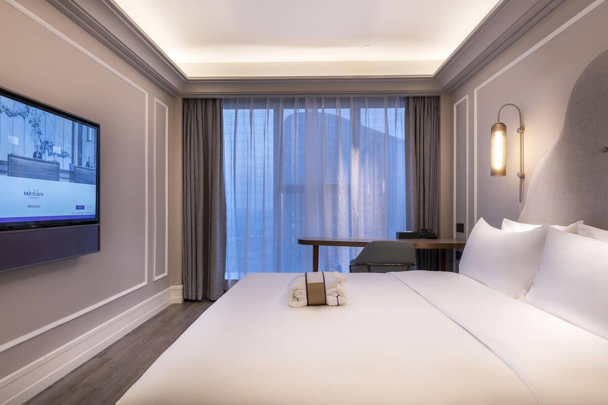 Hotelkamer met een groot bed, een bureau, een stoel, een aan de muur bevestigde televisie en een raam met gordijnen.