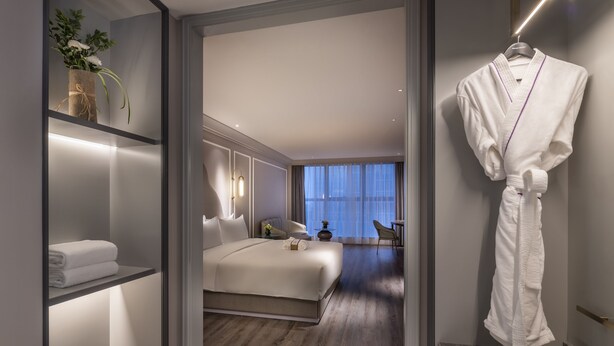 A modern hotel room with a neatly made bed, a robe hanging on the wall, and a view of the city through large windows. - Mercure Suzhou Jinji Lake (Suzhou)