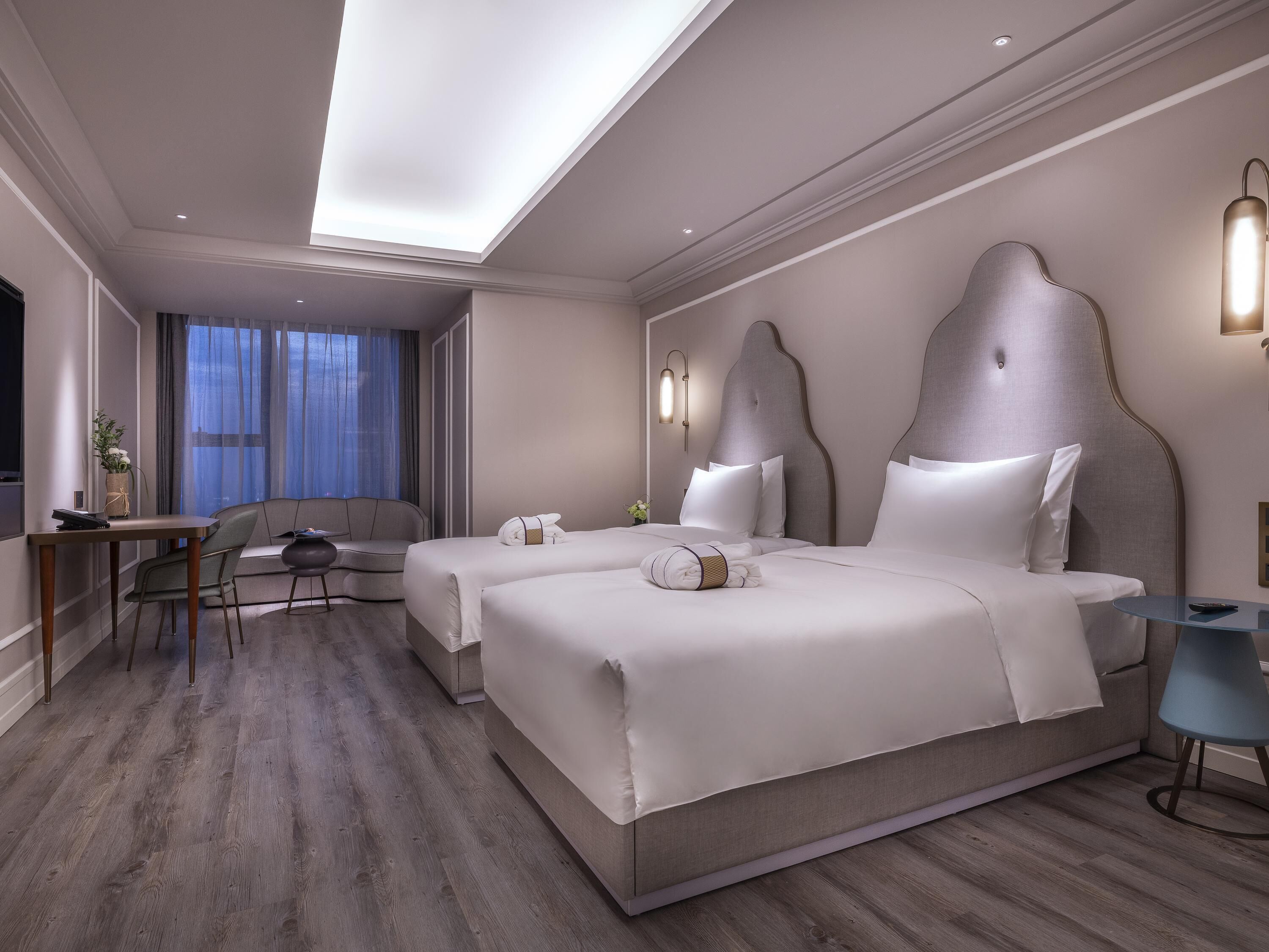 A modern hotel room with two beds, a desk, a chair, and a small table.