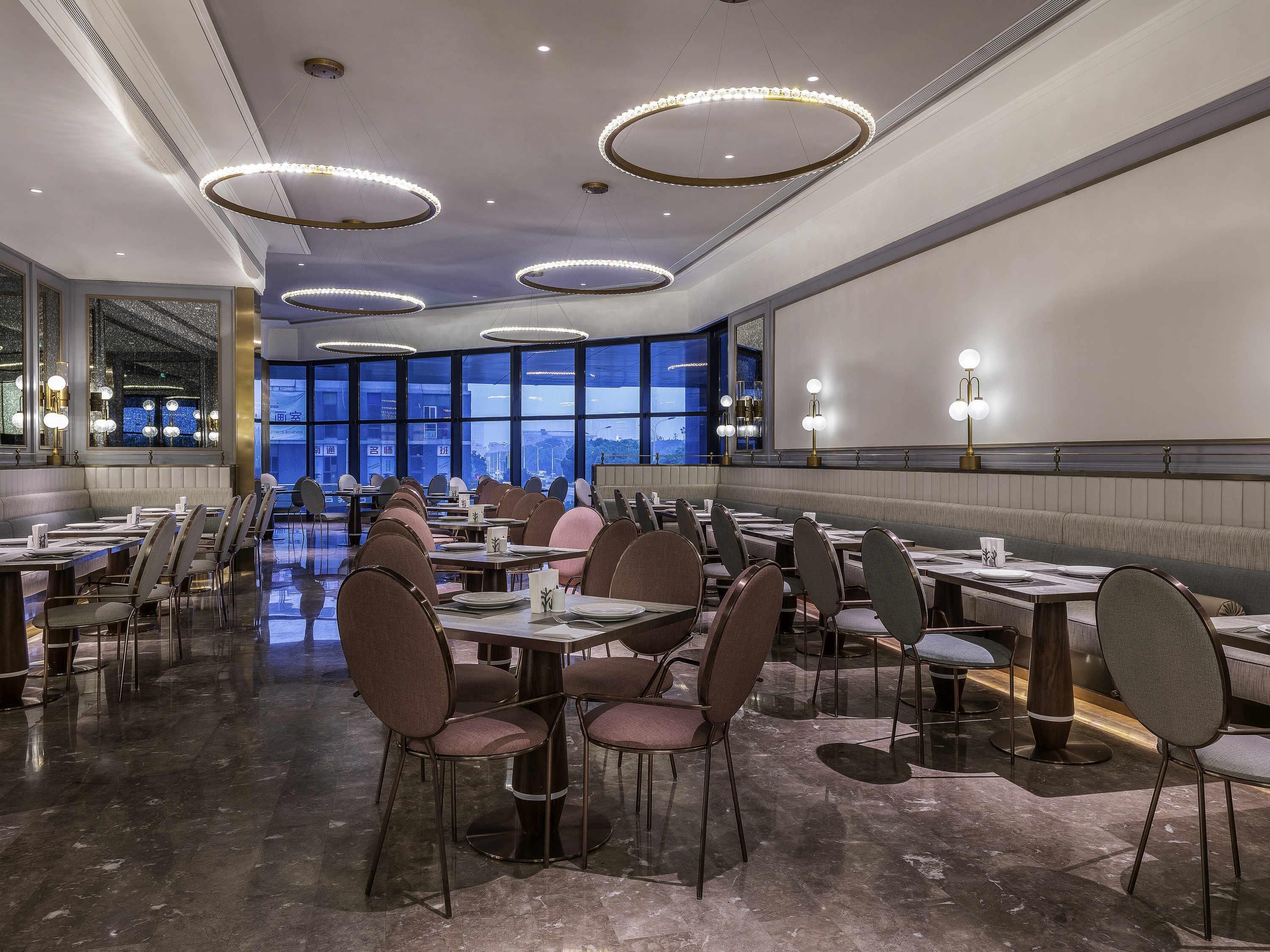 A spacious dining area with multiple tables and chairs, large windows offering a view, and modern circular ceiling lights.
