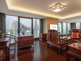 Amplia habitación de hotel con un ventanal grande que ofrece vistas a la ciudad, un escritorio de madera tradicional, un sofá cómodo y un moderno aplique de techo.