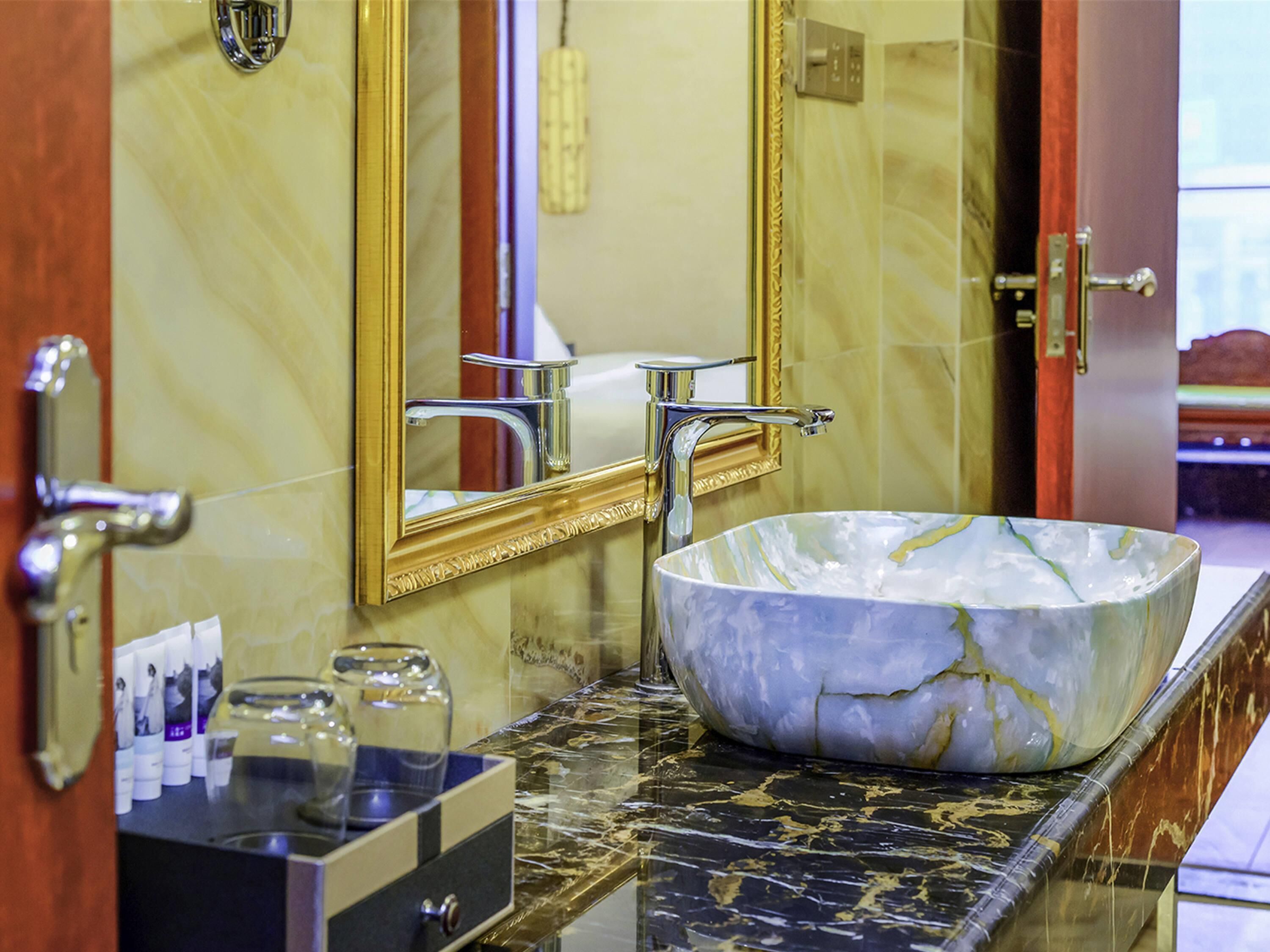 A bathroom with a marble sink, a gold-framed mirror, and a vanity with toiletries.