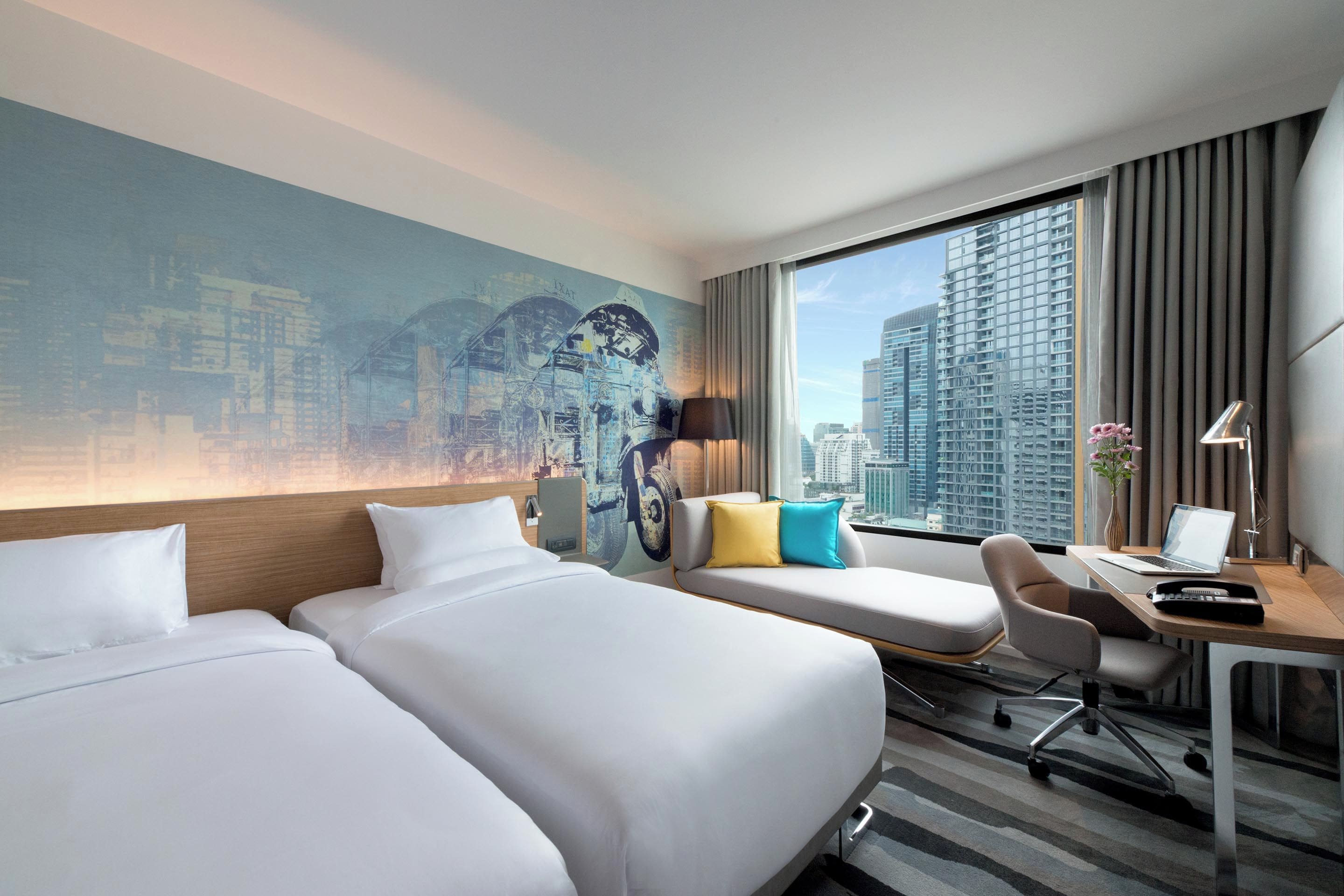 A hotel room with two beds, a desk, a chair, and a large window offering a city view.