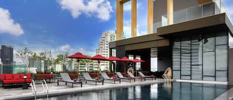 Una piscina en la azotea con sillones y sombrillas, un edificio moderno con ventanas amplias y un cielo azul despejado.