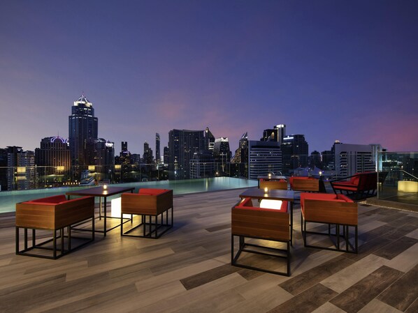 Un bar en la azotea con mobiliario de madera, una piscina y vistas al horizonte urbano al atardecer.