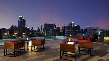 A rooftop bar with wooden furniture, a pool, and a city skyline view at dusk.