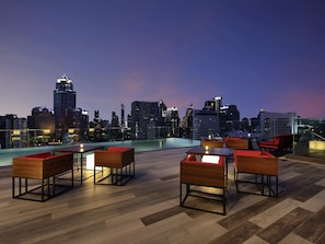 Un bar en la azotea con mobiliario de madera, una piscina y vistas al horizonte urbano al atardecer.