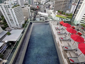 Una piscina en la azotea con sillones de descanso y sombrillas, rodeada de edificios altos.