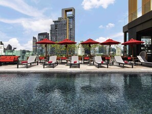 Un espace piscine aménagé sur le toit, avec des chaises longues et des parasols rouges, entouré d’immeubles modernes.