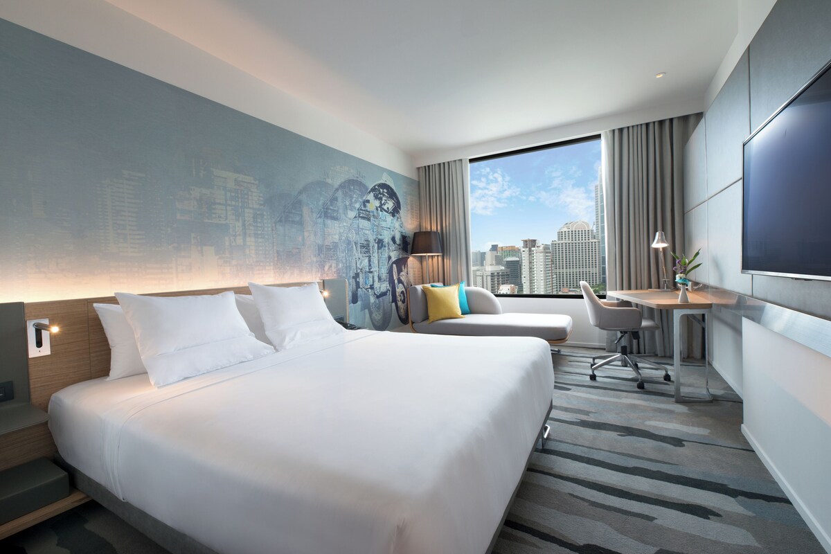 Una habitación de hotel moderna con una cama grande, un escritorio, una silla y vistas a la ciudad.