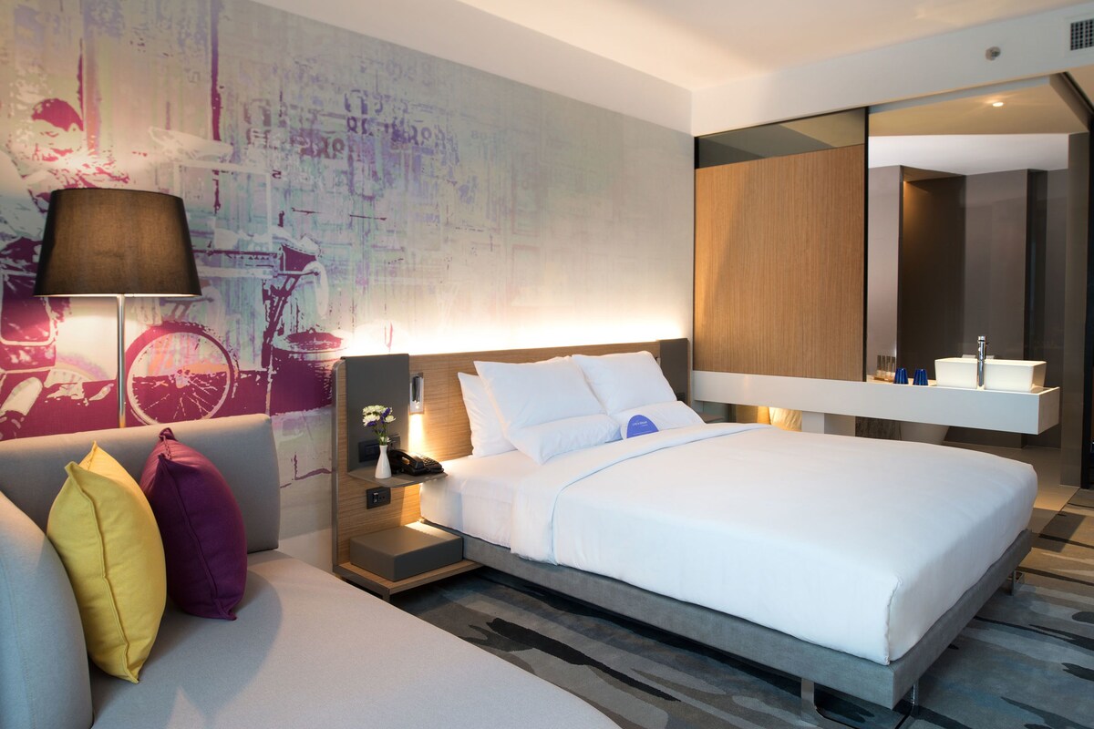 Una habitación de hotel moderna con una cama grande, un sofá con cojines, una mesita de noche con lámpara y un espejo montado en la pared.